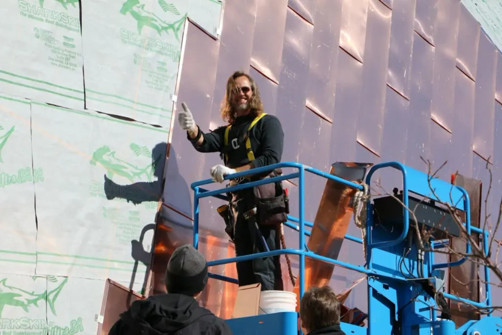 J.R.B. installer on a lift giving a thumbs up in front of a copper wall